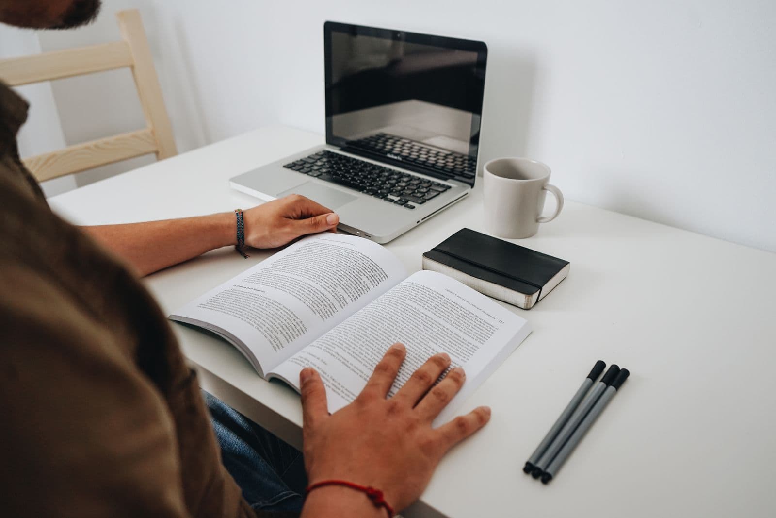 A person reading a book at a desk with a laptop